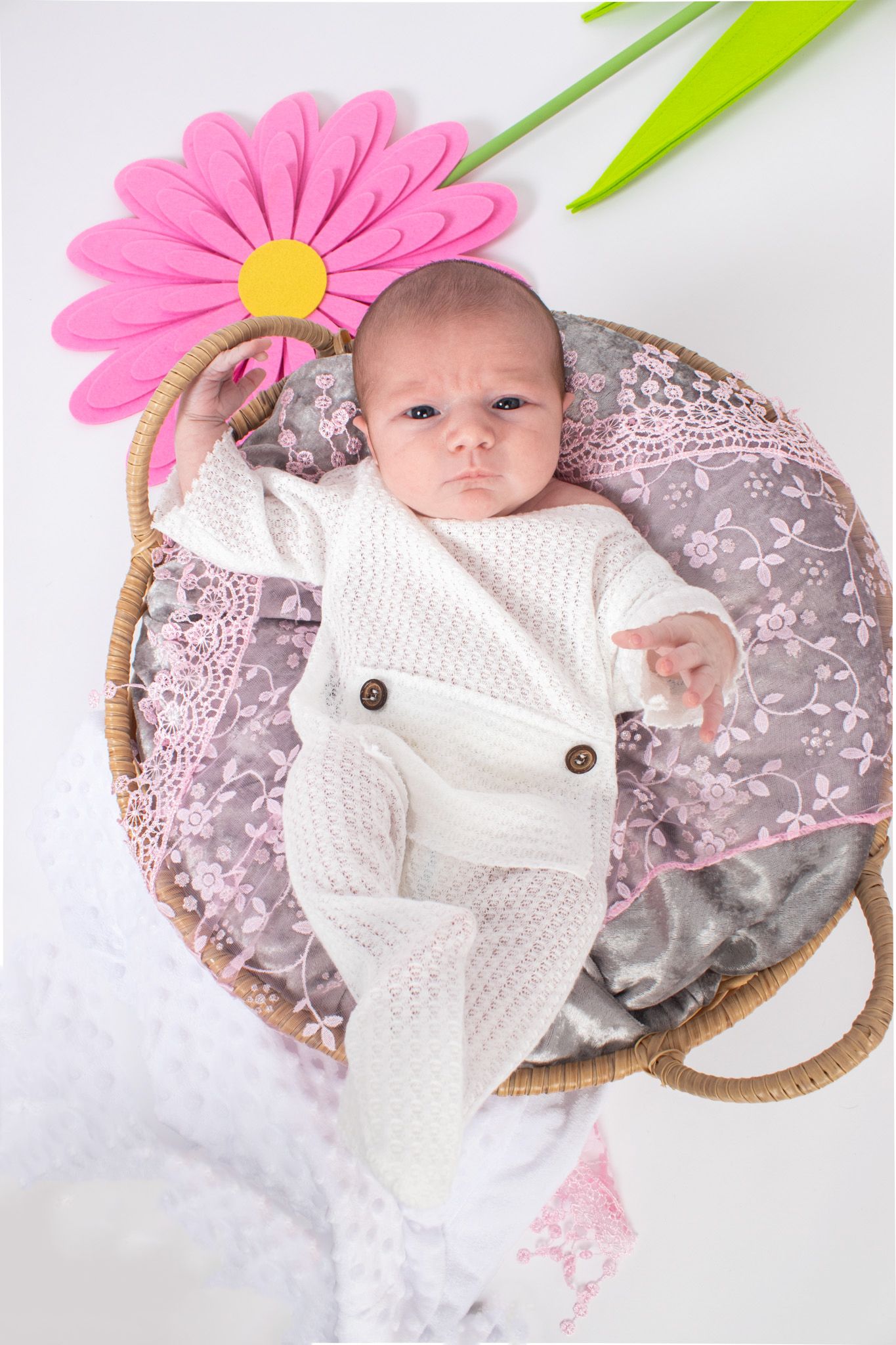 A baby in white knit clothing lies in a basket with a pink flower decoration.