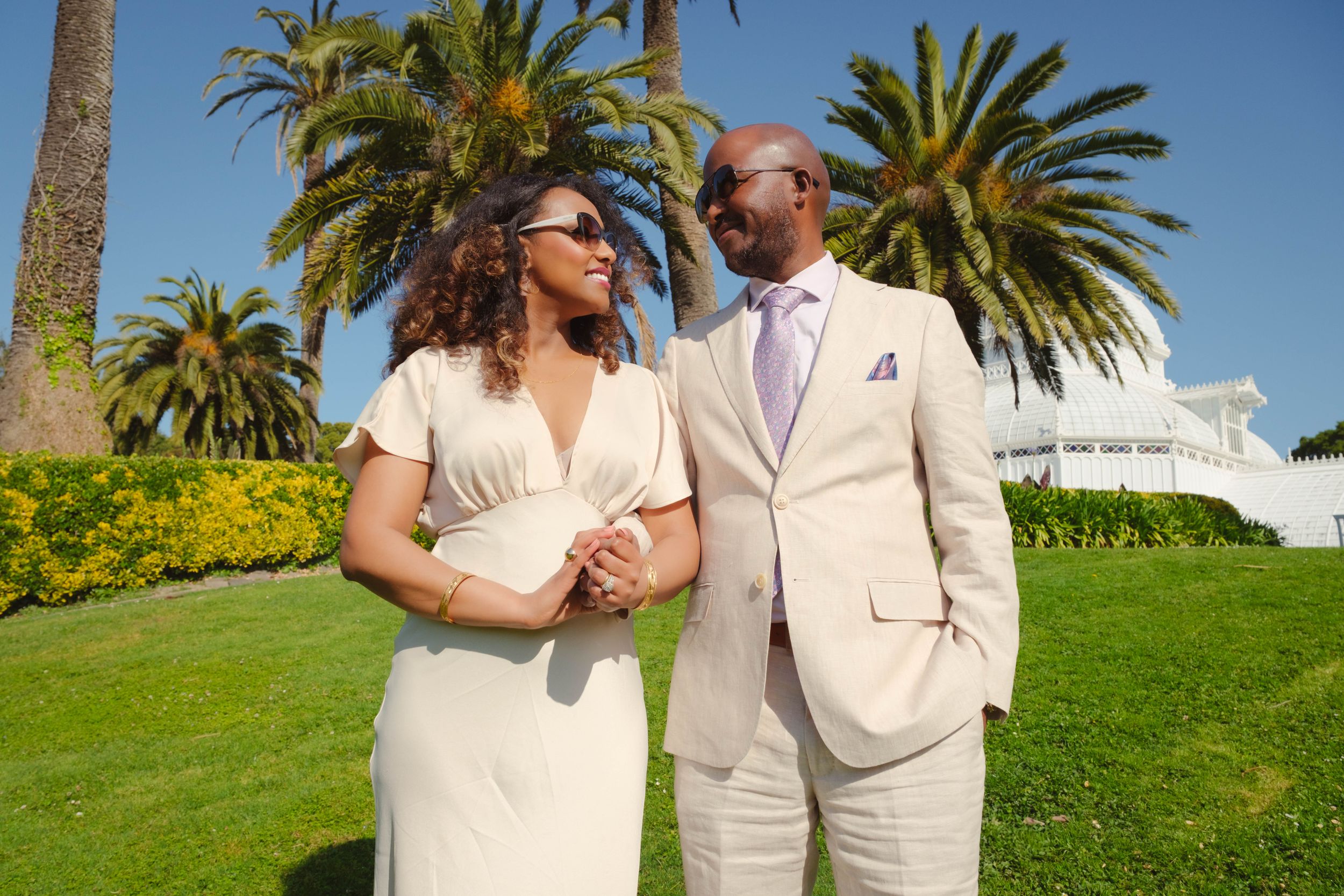 A couple in cream-colored wedding attire embraces on a grassy hill with palm trees and blue sky in the background.