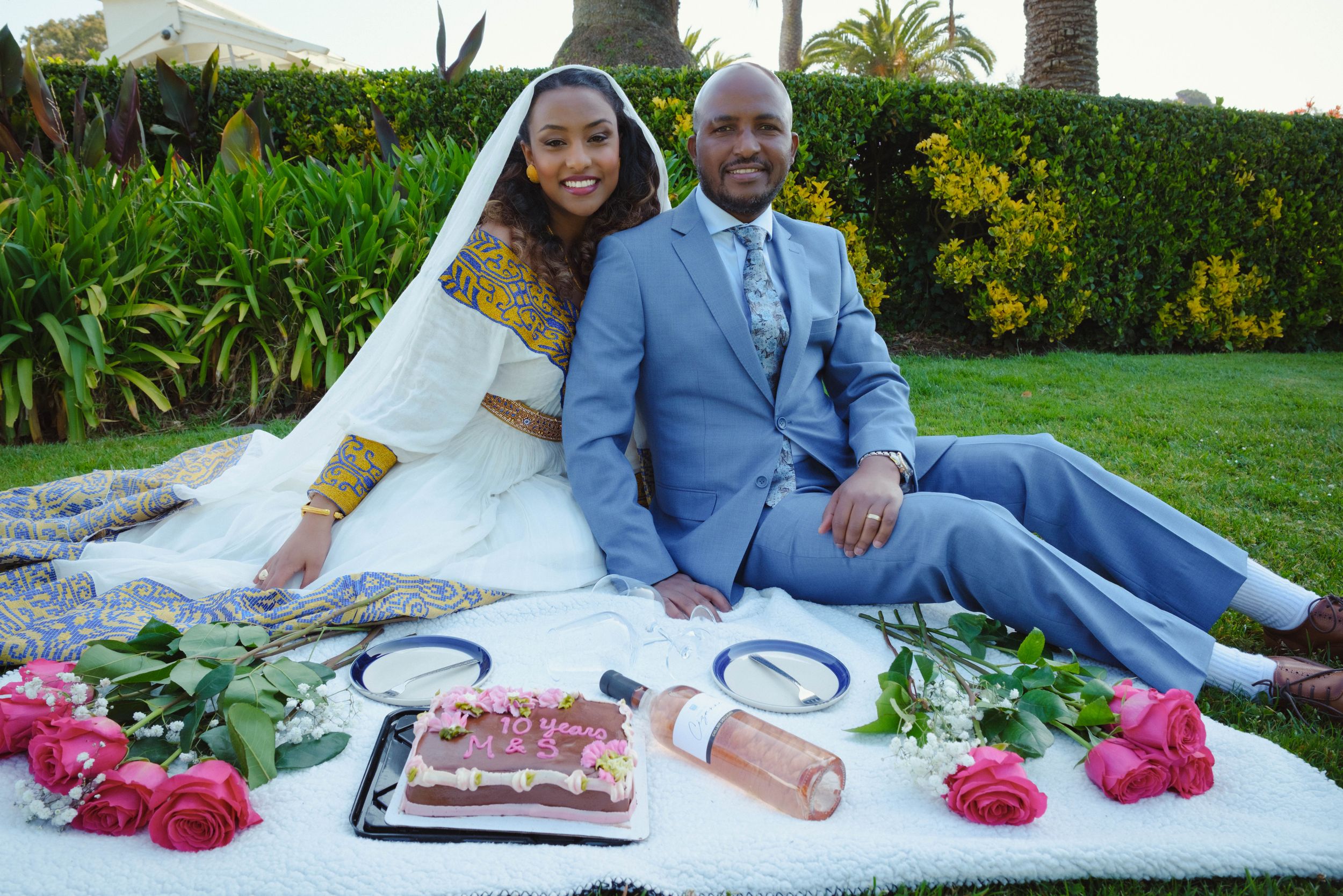 A couple in wedding attire sits on a white blanket with pink flowers during an outdoor garden photo session.