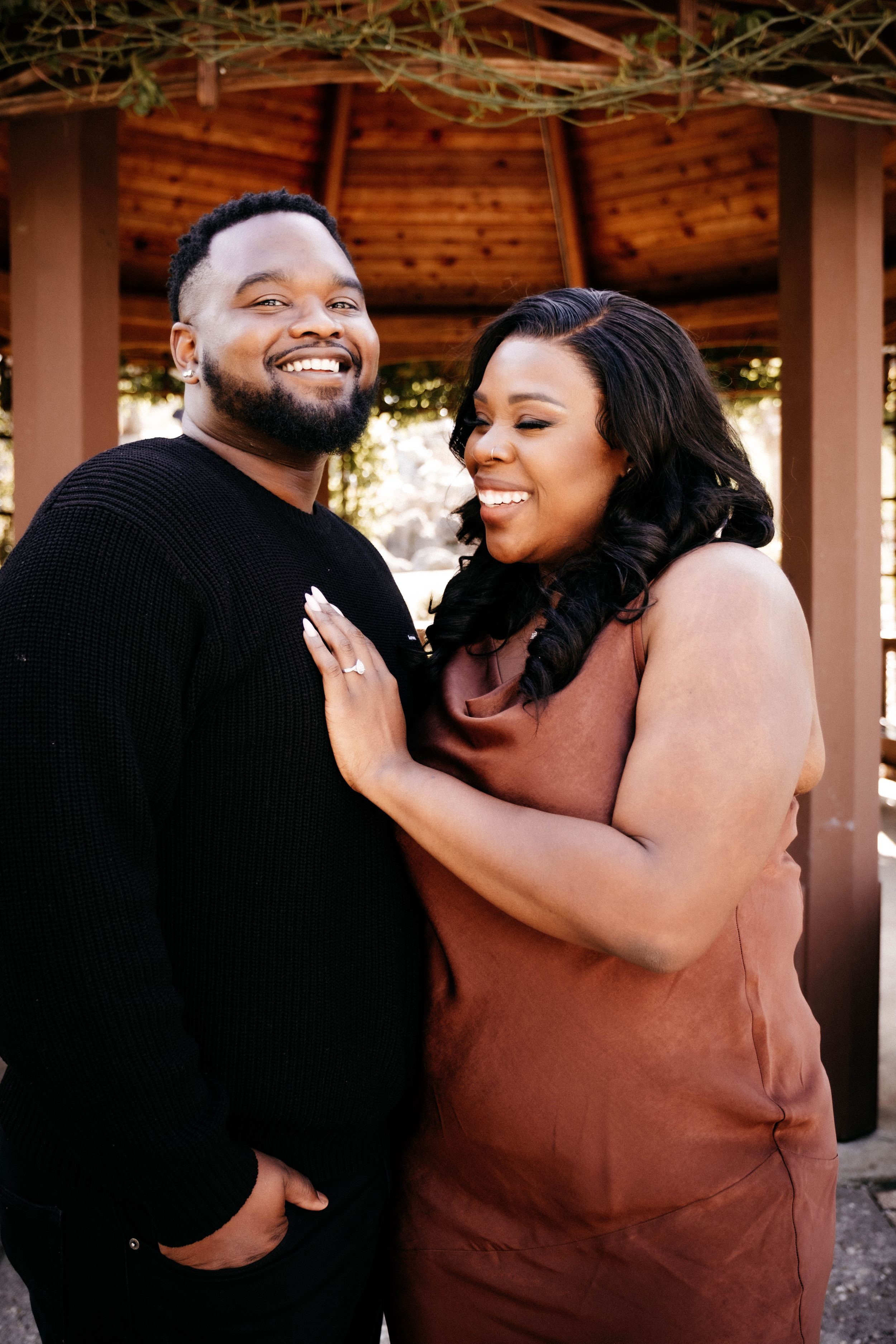 A couple shares joyful laughter and embraces during an outdoor photoshoot under a wooden pergola.