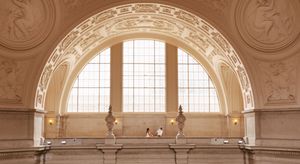 Elegant neoclassical hall with ornate arched ceiling, large arched window, and decorative sculptures in a grand interior space.