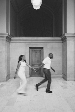 Black and white photo of a couple holding hands and running through an elegant building with arched ceilings.