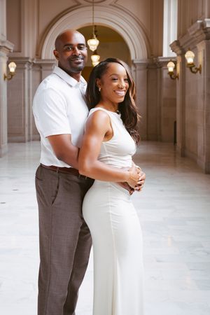 A smiling couple poses elegantly in a grand marble hallway, the woman in white dress, man in white shirt and brown pants.