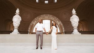 Couple holding hands in elegant San Francisco City Hall rotunda, woman in white gown, man in white shirt and gray pants.