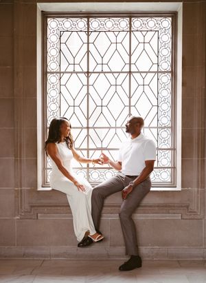 Couple in elegant attire sitting together by an ornate leaded glass window, sharing an intimate moment.