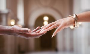 Two hands gently reaching toward each other in an elegant hall, one adorned with a bracelet and ring.