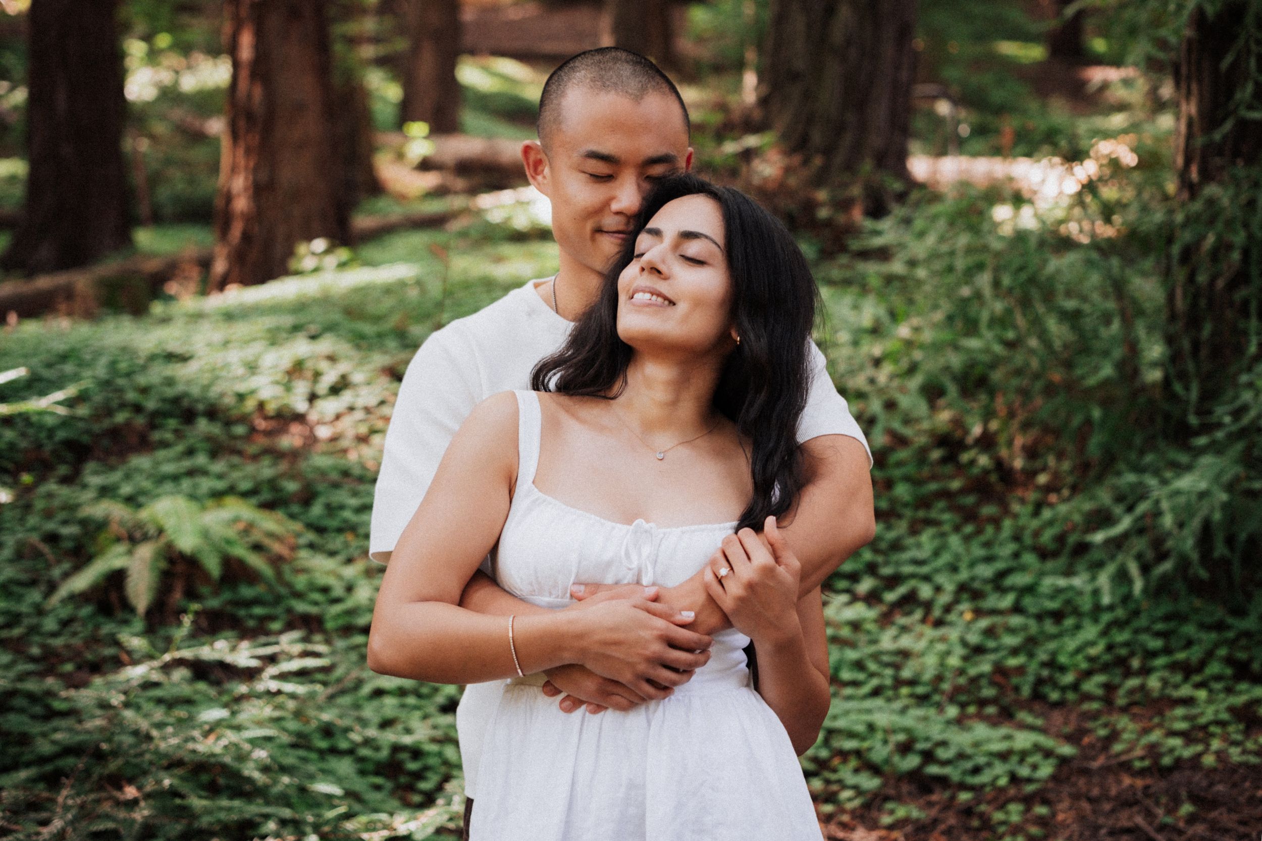 Couple embracing in a lush green forest, woman in white dress smiling with eyes closed, man hugging her from behind.