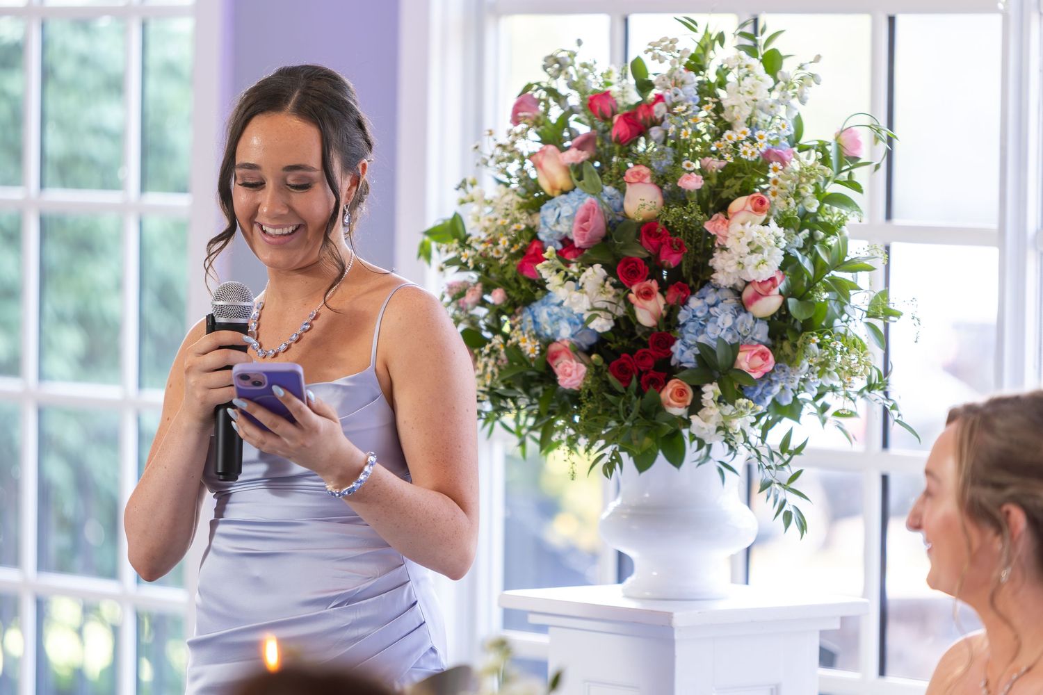 Wedding guests laugh and socialize during an elegant reception with floral arrangements and bright windows.