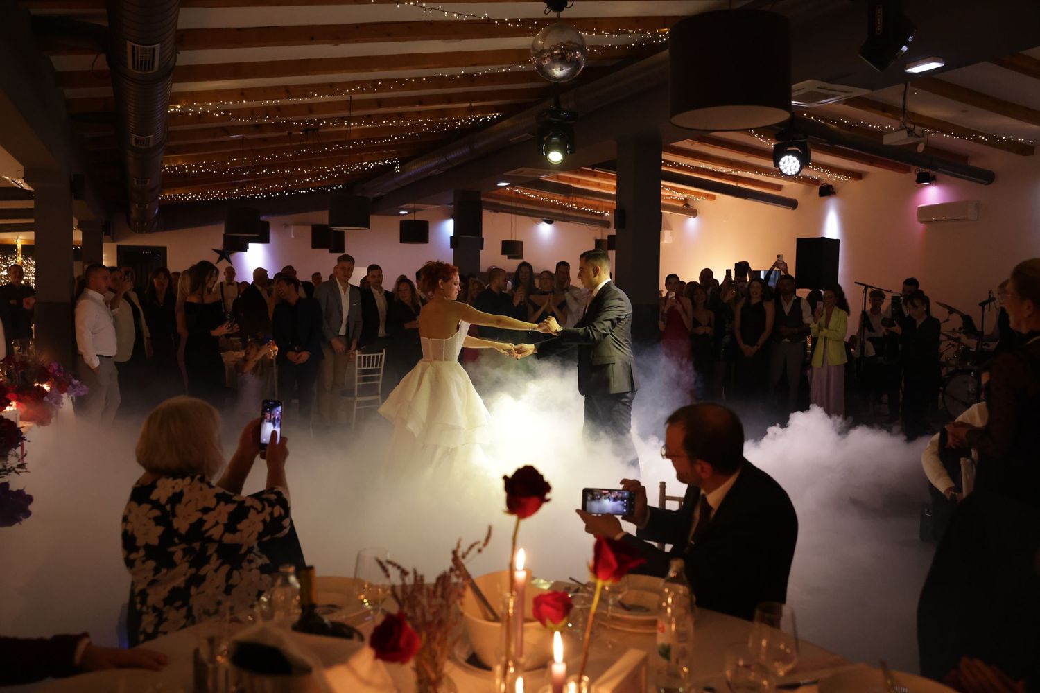 A couple shares their first dance at a wedding reception with fog effects and red floral decorations in a rustic venue.