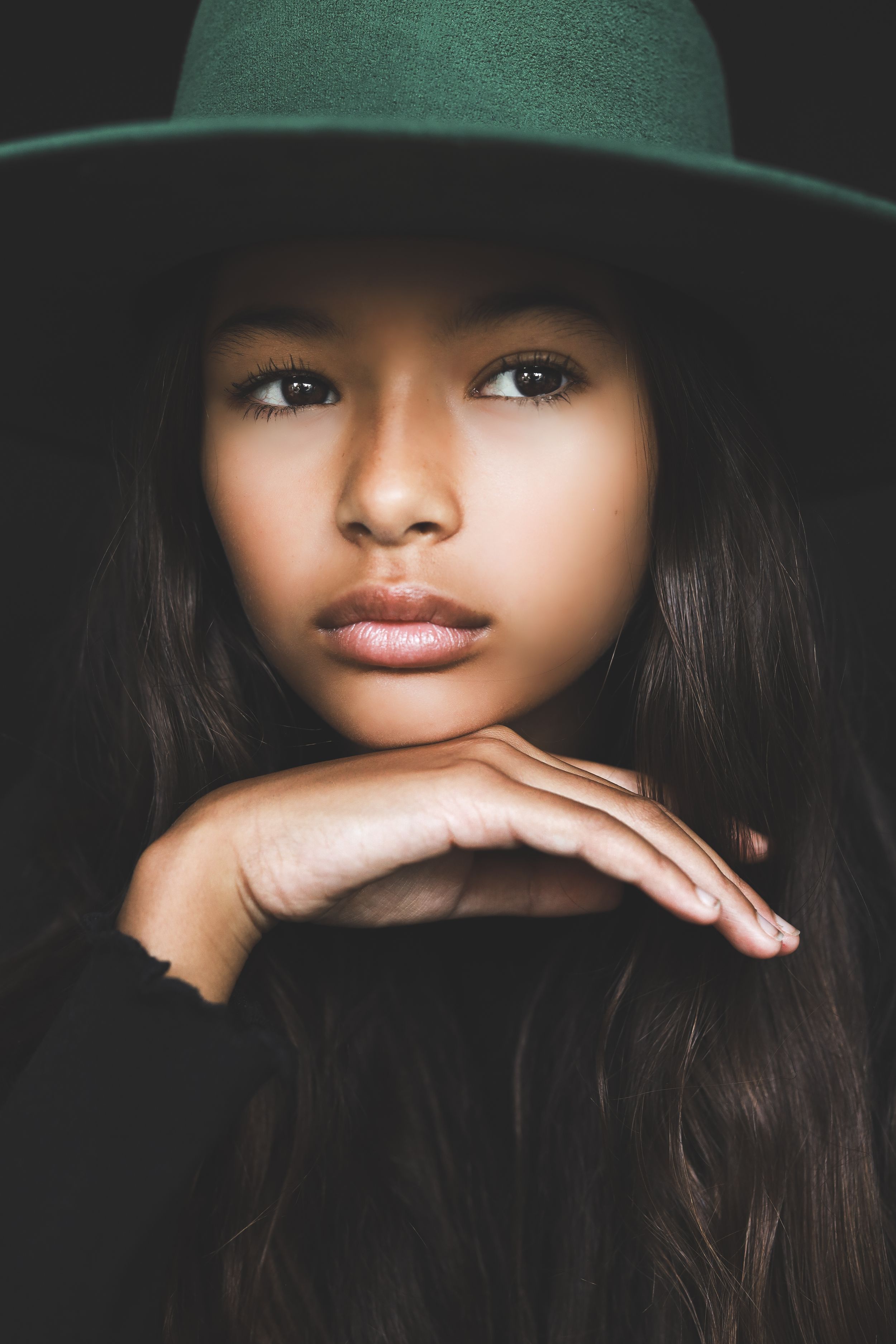 Young woman with long hair wearing a wide-brim green felt hat, resting chin on hands against dark background.