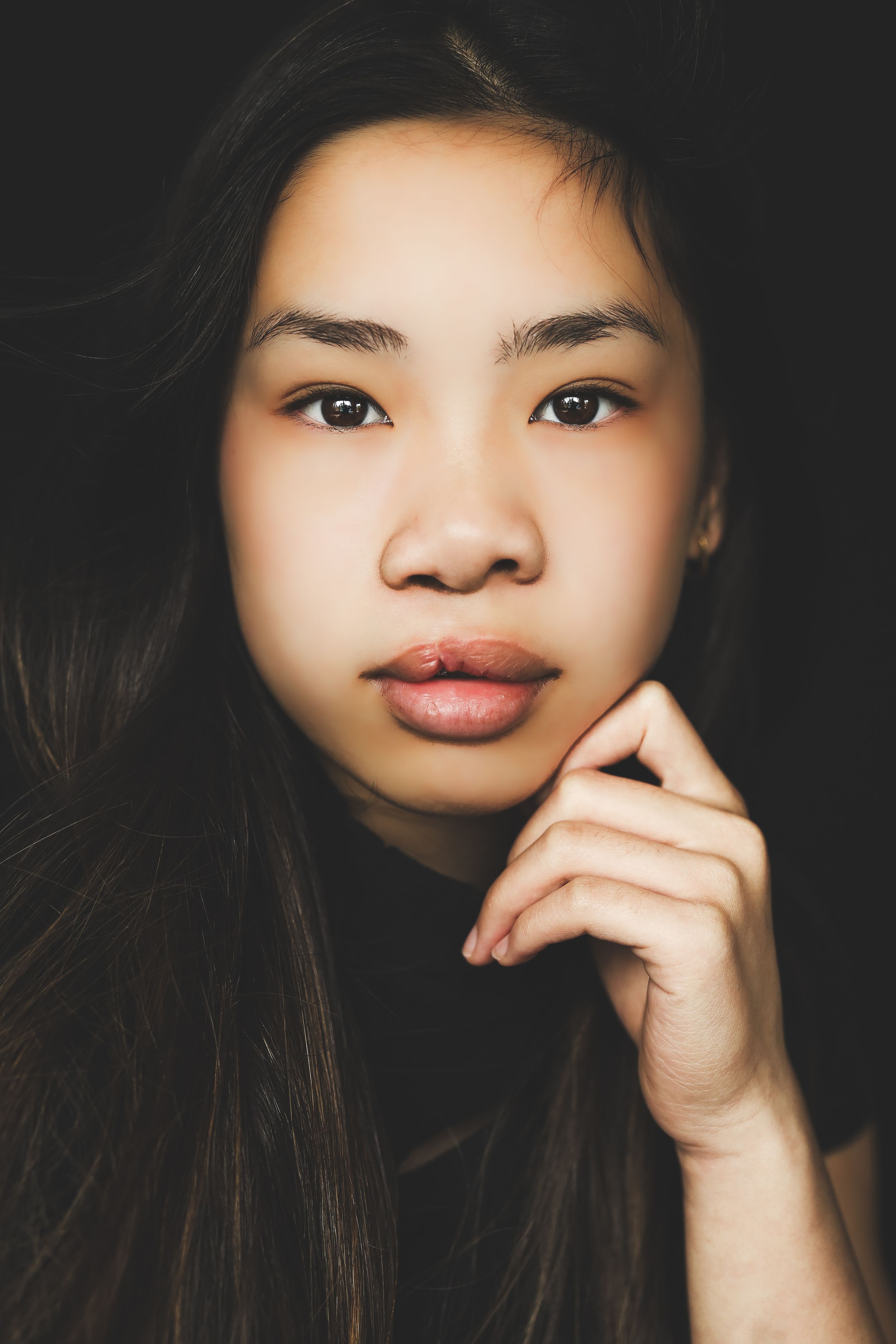 Young Asian woman with long dark hair resting chin on hand, gazing at camera against dark background.