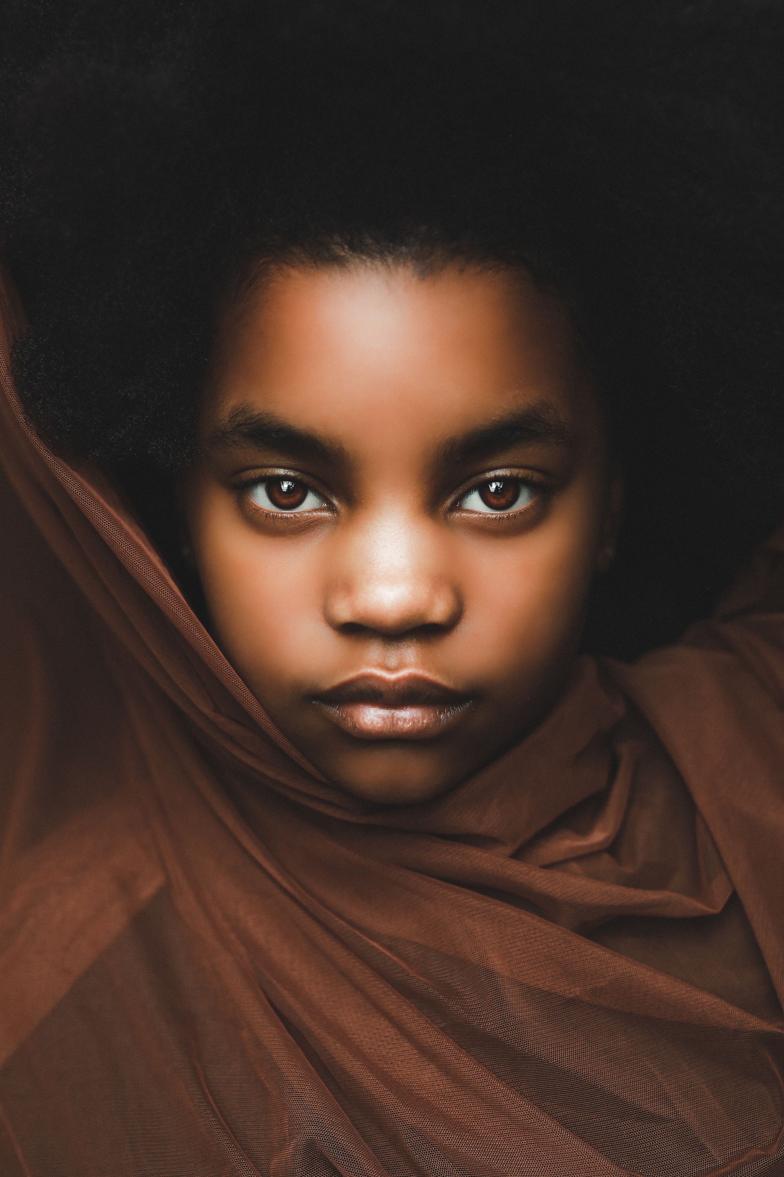 Portrait of a young Black girl with an afro, wrapped in brown fabric, gazing intensely at the camera.