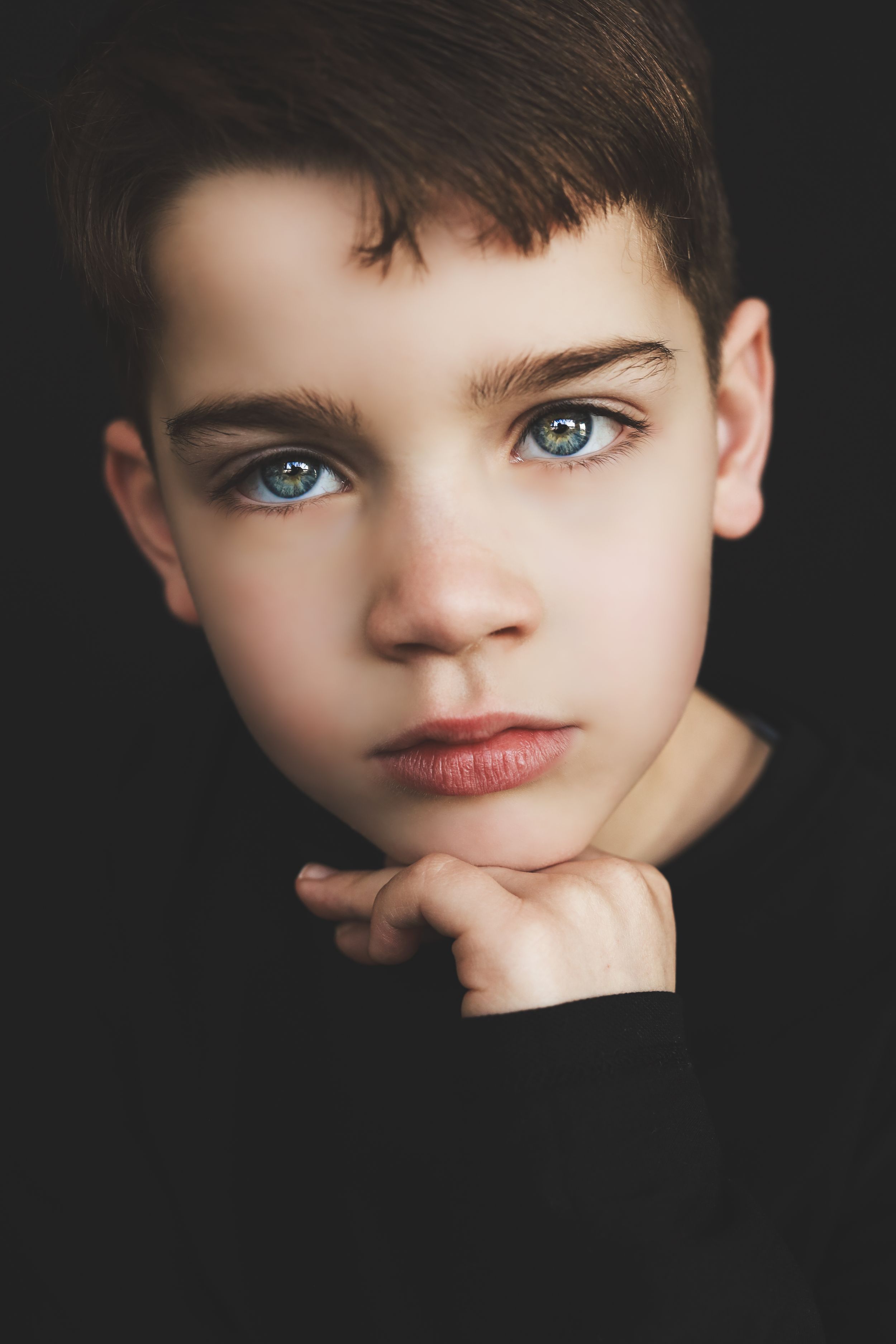 Portrait of a young boy with striking blue-green eyes and dark hair, resting chin on hand against dark background.