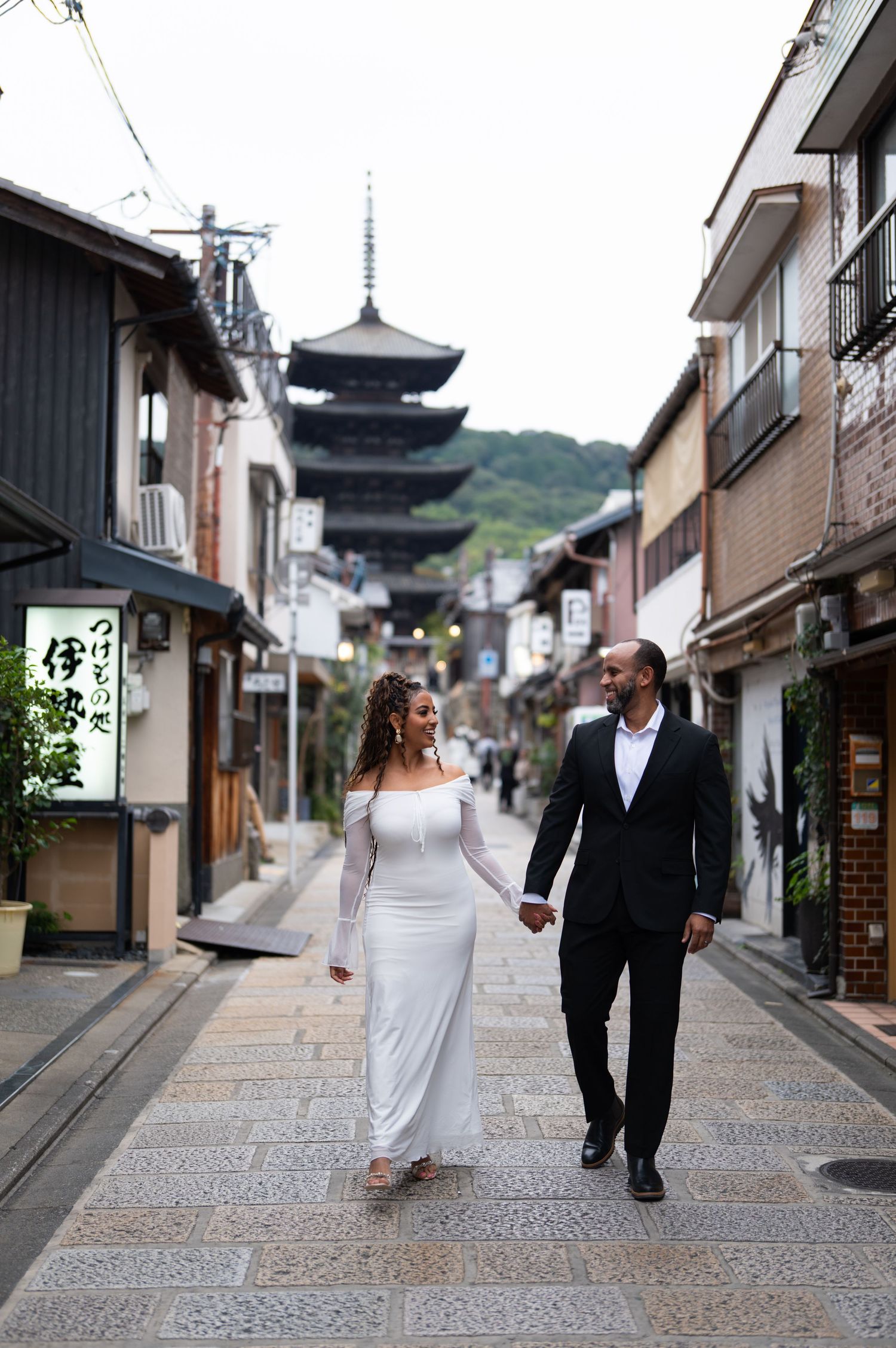 Couple walks hand-in-hand down Kyoto street with Yasaka Pagoda in background, woman in white dress.