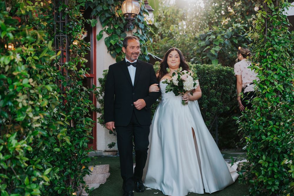 Wedding couple walks through a lush garden pathway surrounded by green foliage and twinkling lights.