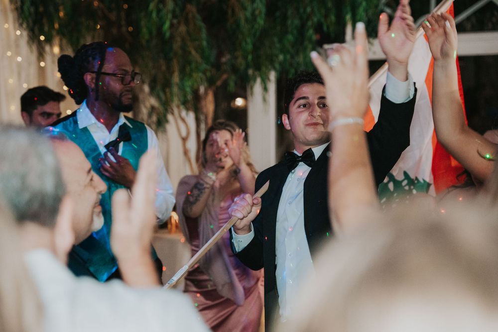 Wedding celebration with guests raising hands and dancing in a dimly lit venue with decorative greenery overhead.