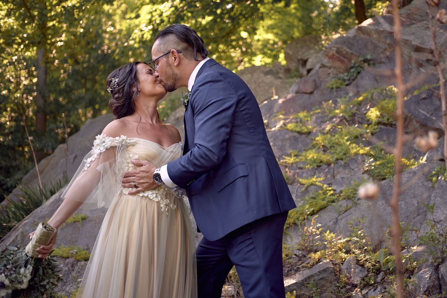 A couple in formal attire shares romantic moments in a wooded outdoor setting during a wedding photoshoot.