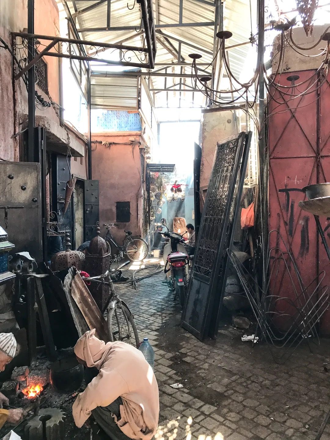 A narrow alley with craftsmen working on metal goods beside rust-coloured doors and tools.