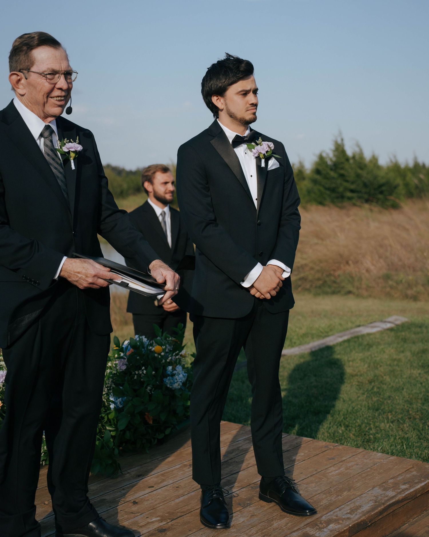 Two men in formal black suits stand on a wooden platform during an outdoor wedding ceremony.
