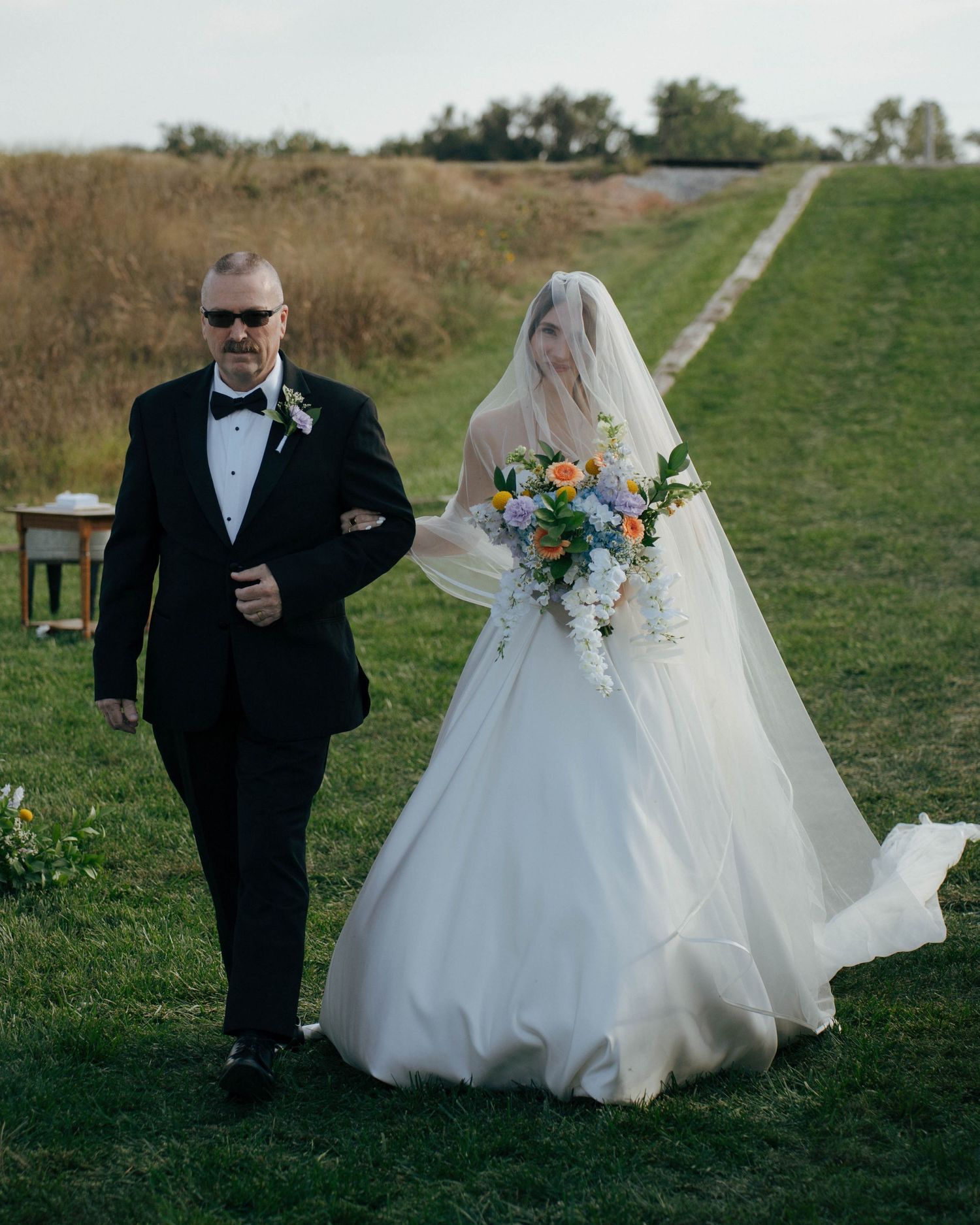 A wedding ceremony on a grassy hillside with a bride in white dress carrying a colorful bouquet walking with an escort.