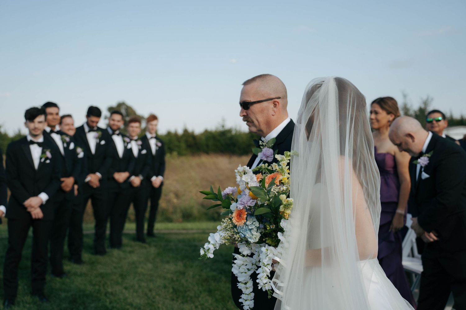 Wedding party gathers outdoors on a grassy hillside during sunset for formal ceremony and group photos.