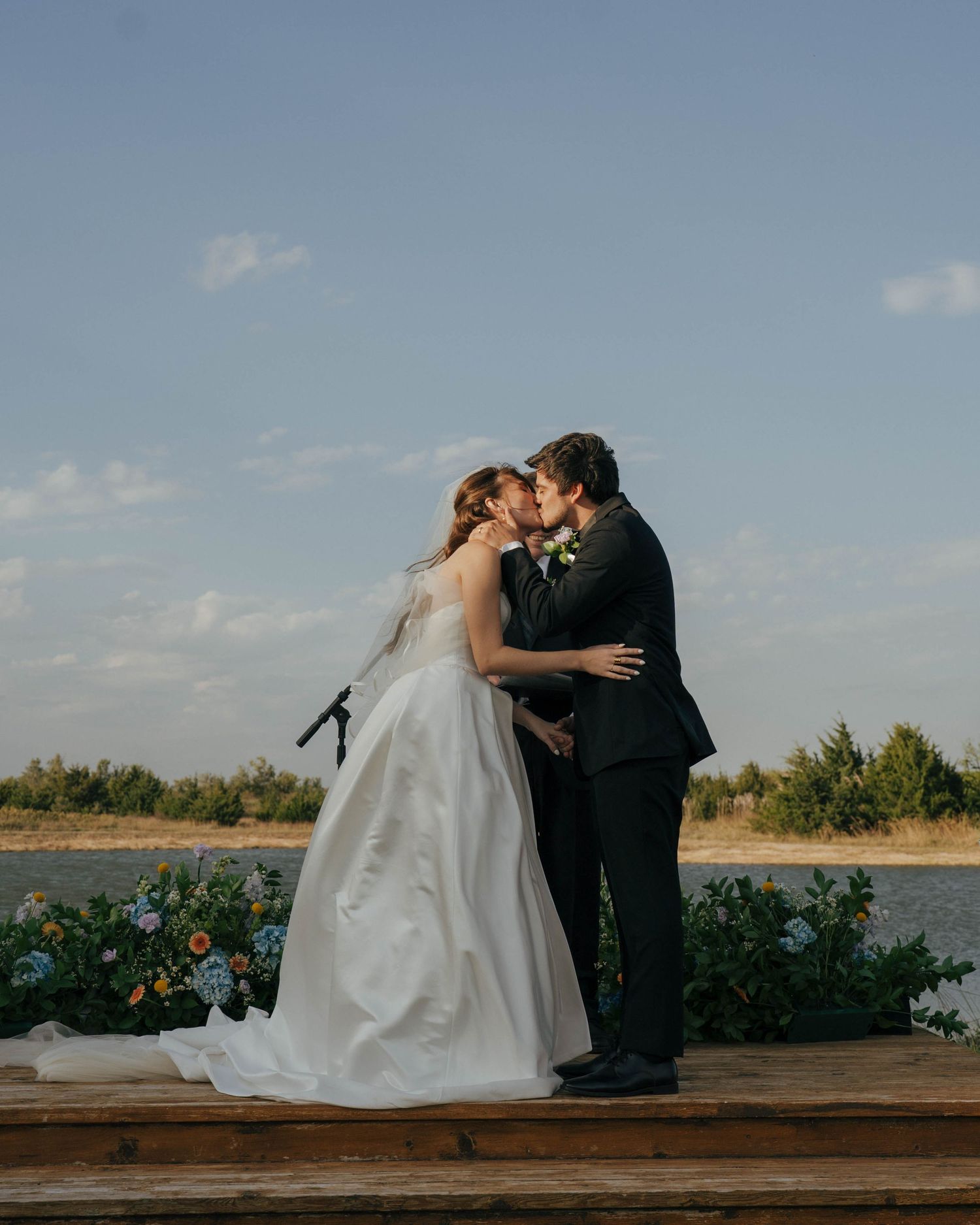 A romantic wedding couple shares tender moments on a rustic wooden deck overlooking a scenic lake at sunset.