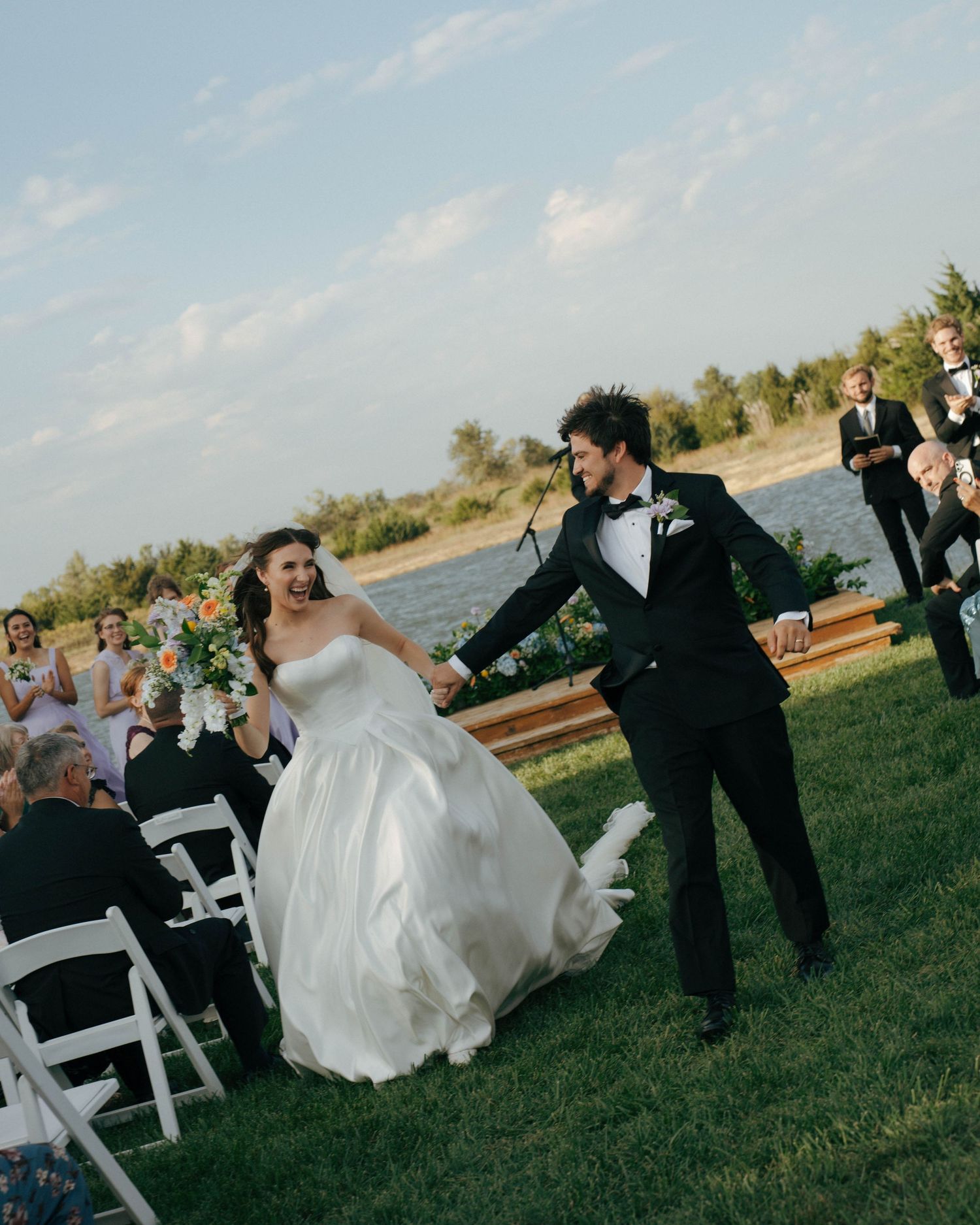 A joyful wedding celebration takes place on a grassy lawn with white chairs and a scenic waterfront backdrop.
