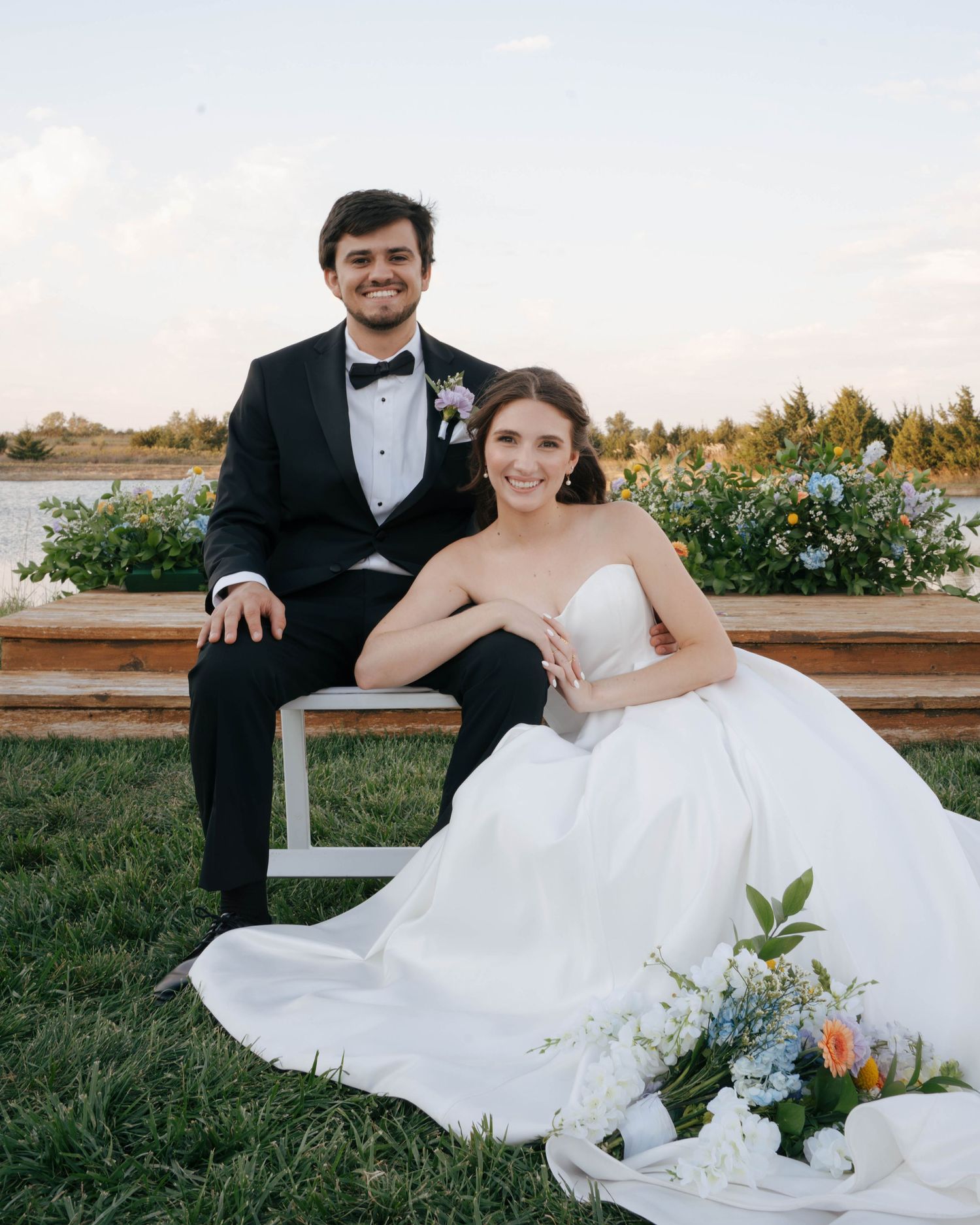 A newlywed couple sits on a bench by a waterfront garden during their wedding photoshoot.