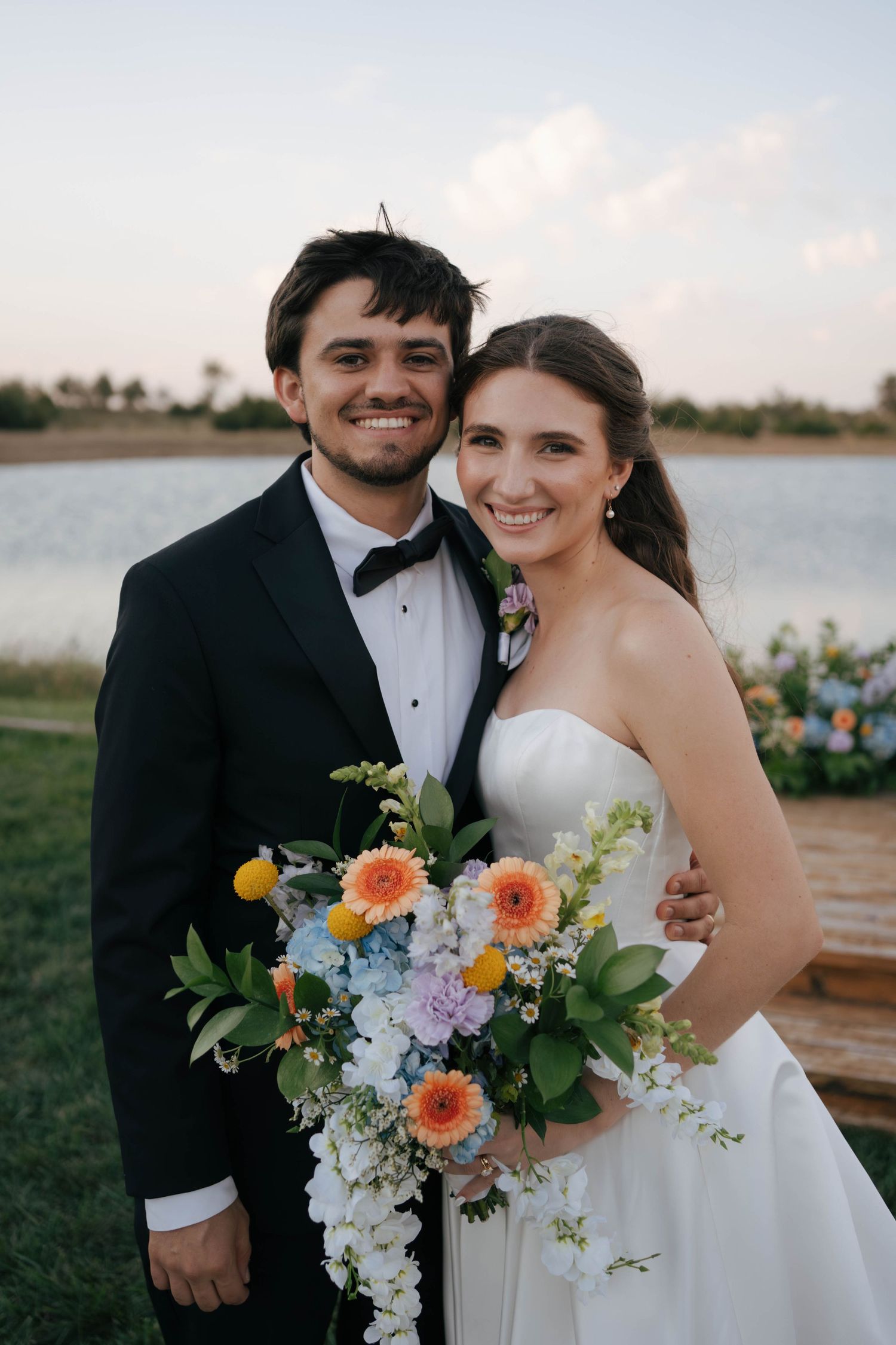 A wedding couple poses by a lake at sunset with a colorful bouquet of coral and yellow flowers.