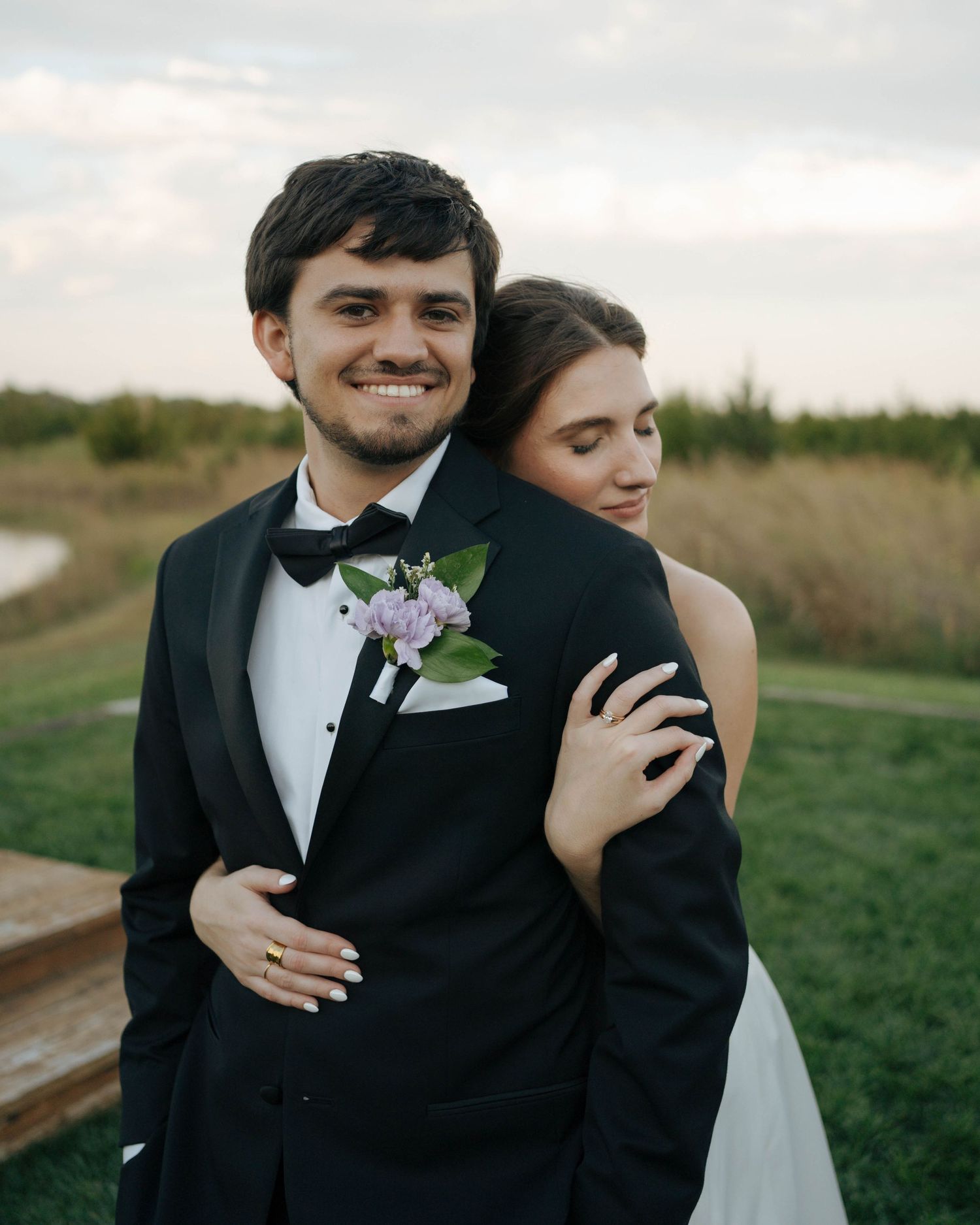 A romantic wedding portrait of a couple in formal attire embracing in a grassy outdoor setting at sunset.