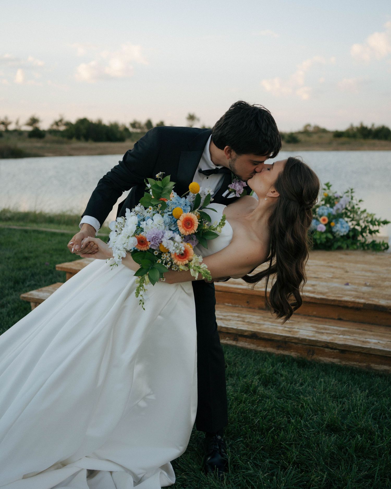 A couple shares a romantic kiss by the water while holding a vibrant bouquet of blue and orange flowers at sunset.