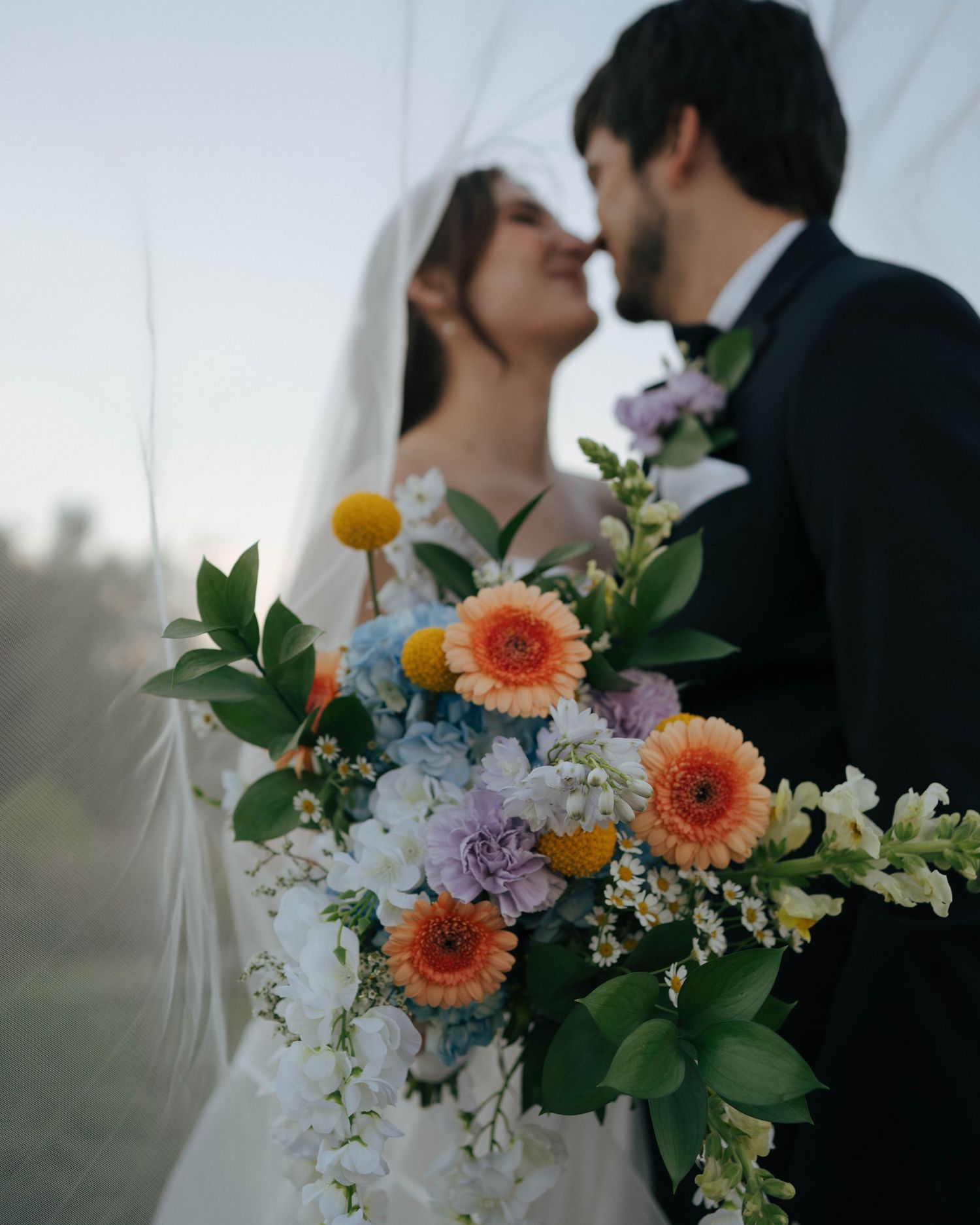 A romantic wedding bouquet featuring coral gerbera daisies, purple flowers, and white blooms with greenery.