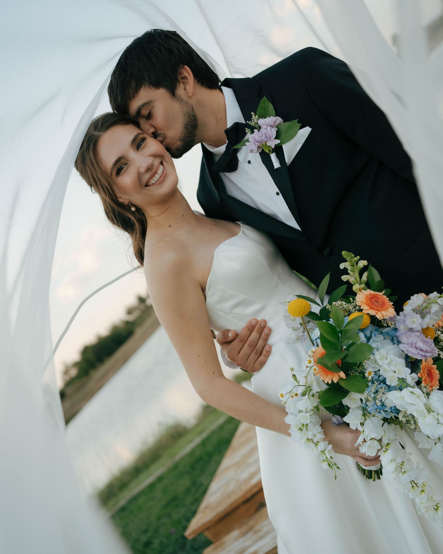Newlywed couple embracing at outdoor wedding ceremony with colorful wildflower bouquet against natural backdrop.