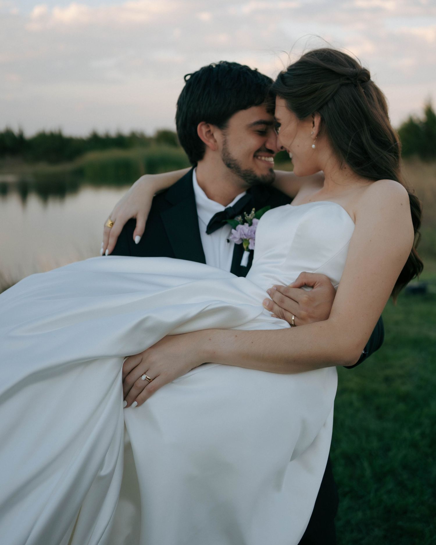 A romantic wedding moment captured by a lake as a couple embraces in formal attire at sunset.