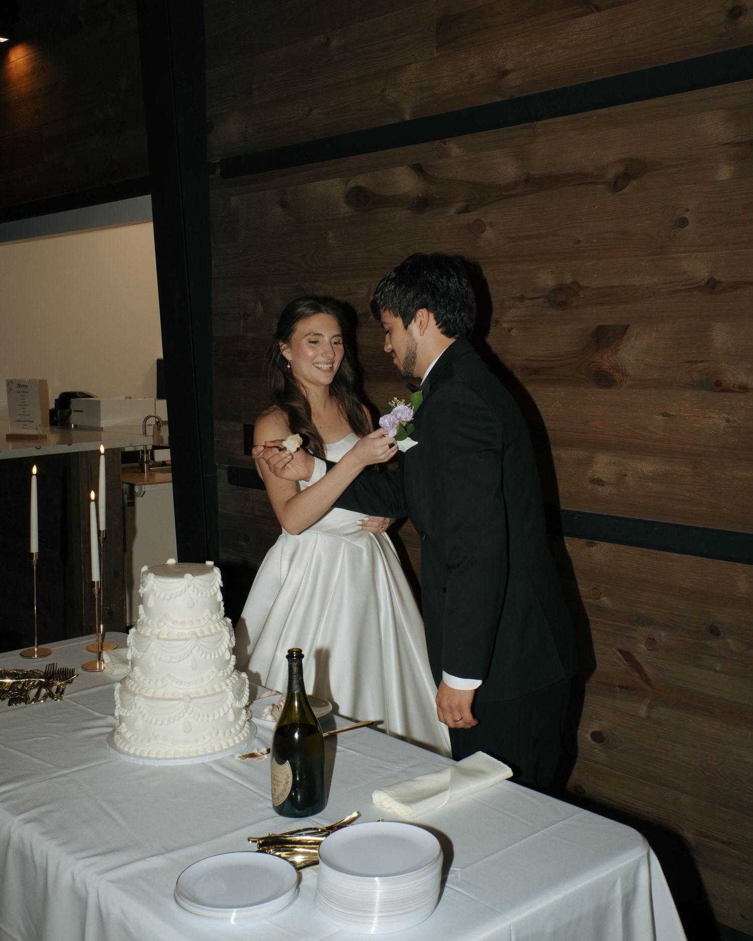 A couple cuts their white wedding cake at a reception with rustic wood paneled walls in the background.