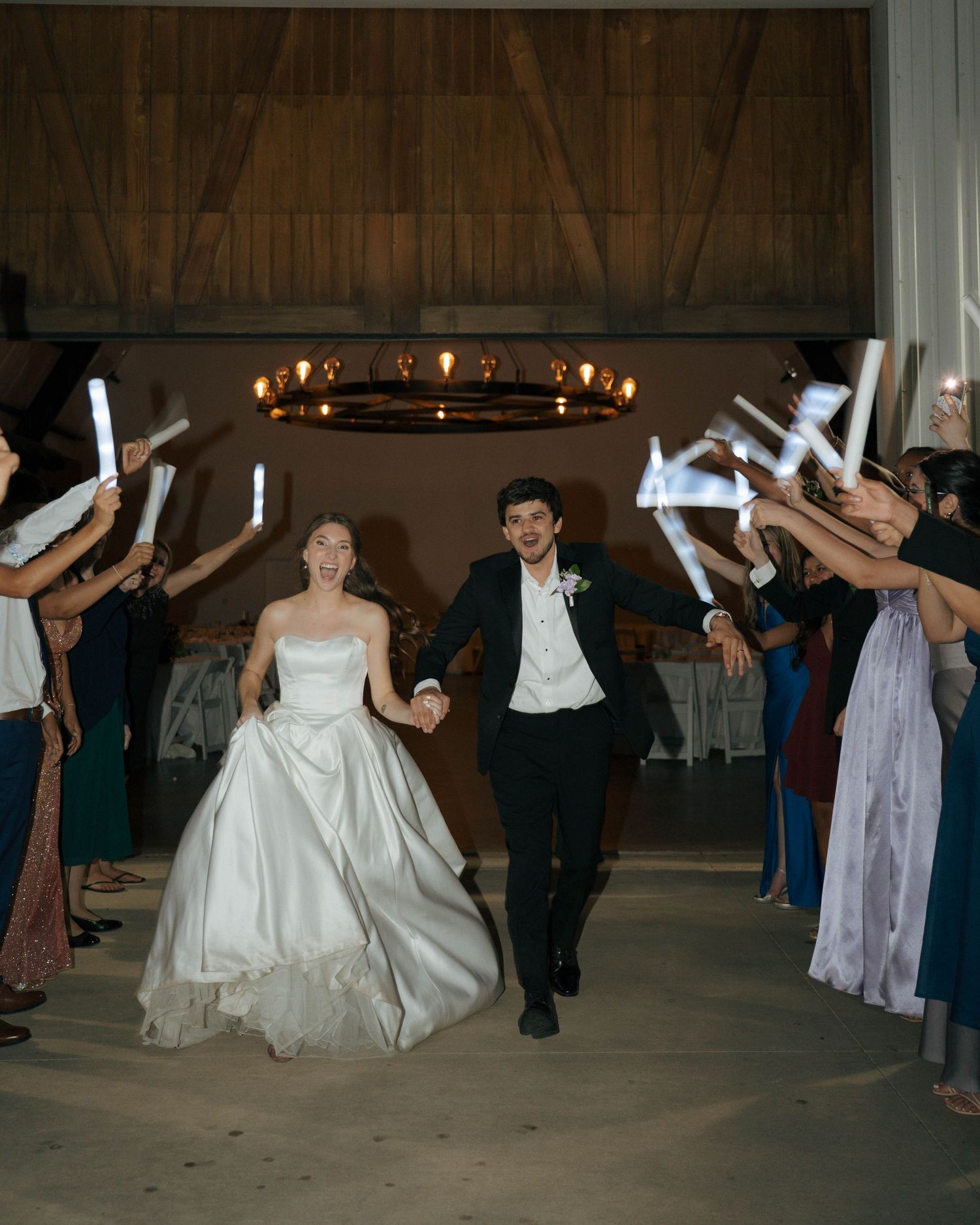Wedding guests dance in a synchronized line formation at a rustic barn reception venue with wooden beams and chandeliers.