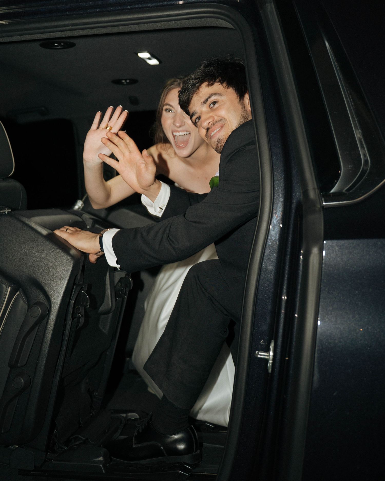 Wedding party members waving from inside a dark vehicle at night.