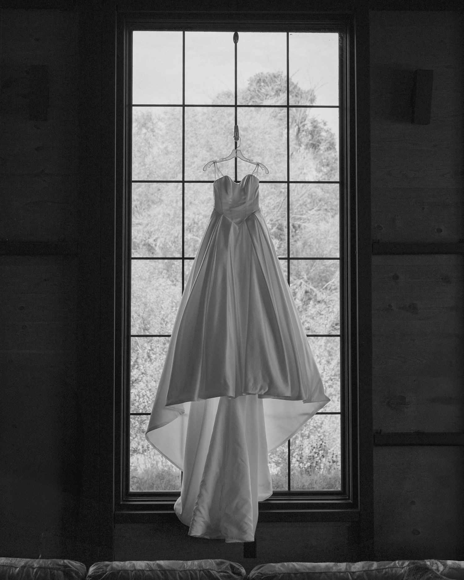 A white wedding dress hangs elegantly in silhouette against a bright window in a dark room.