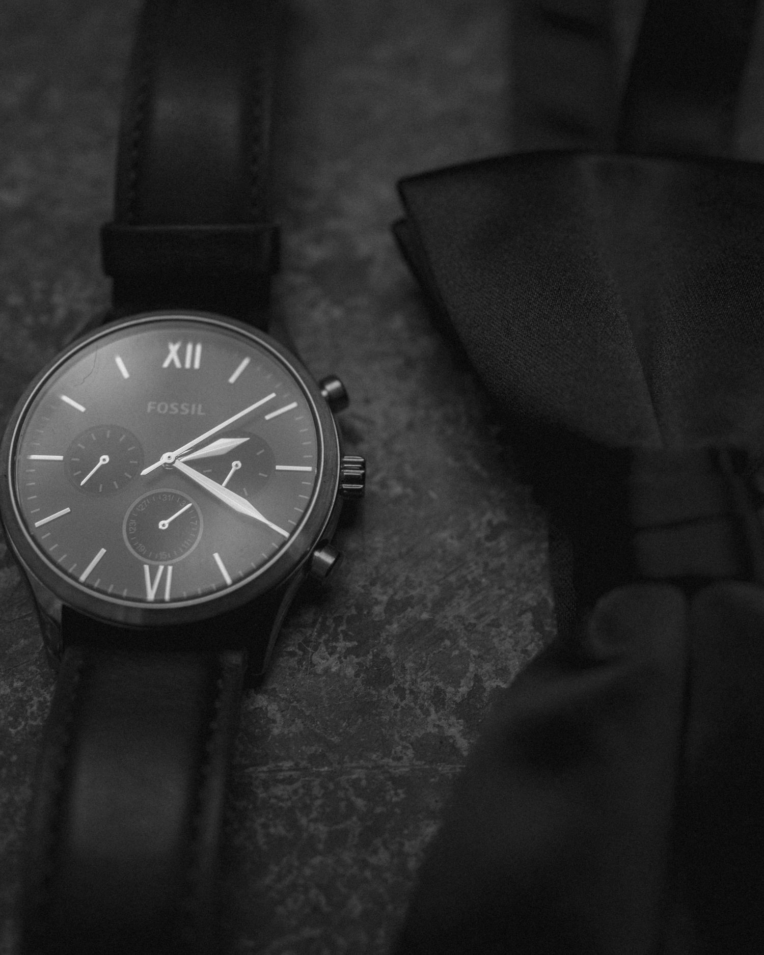 A sleek black chronograph watch with Roman numerals displayed next to a dark bow tie.