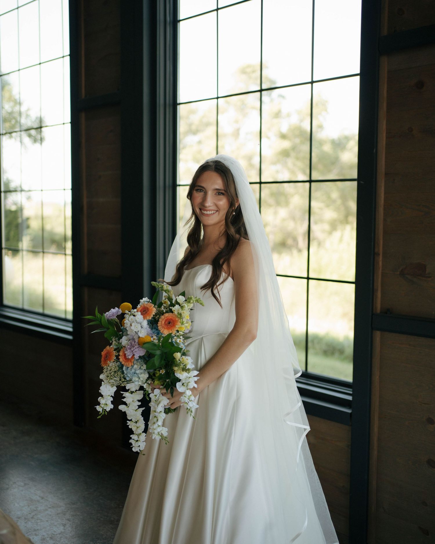 A wedding dress with a flowing train and veil captured in front of large windows in natural light.