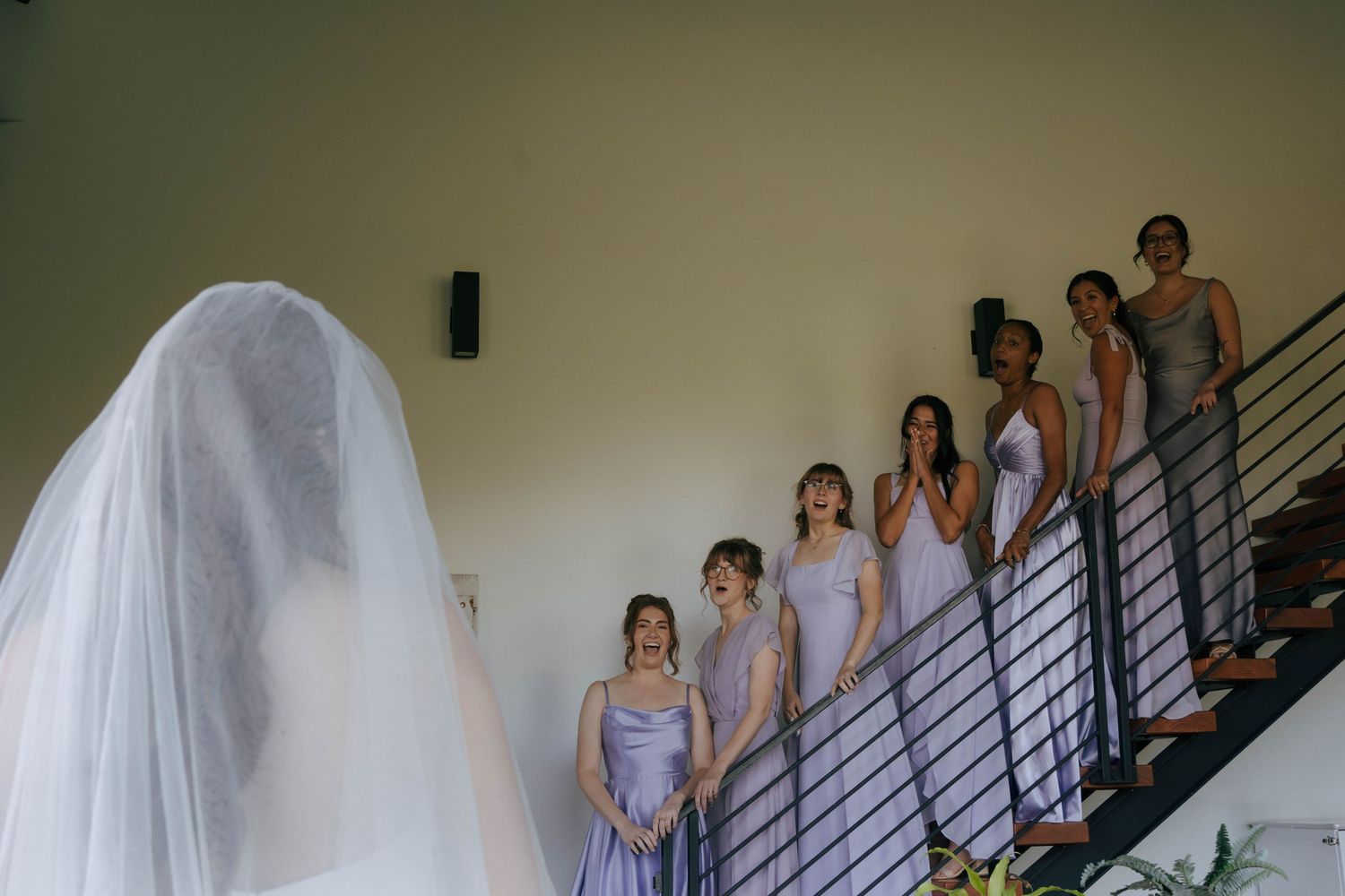 A wedding party descends a modern staircase with bridesmaids in lavender dresses and a bride in white with a flowing veil.