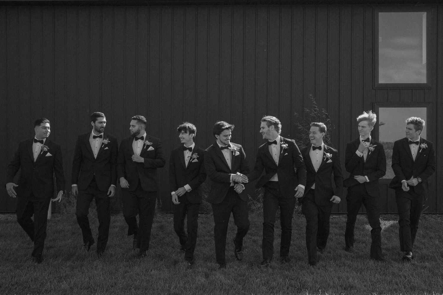 A line of groomsmen in black tuxedos pose together against a dark background for a formal wedding photo.
