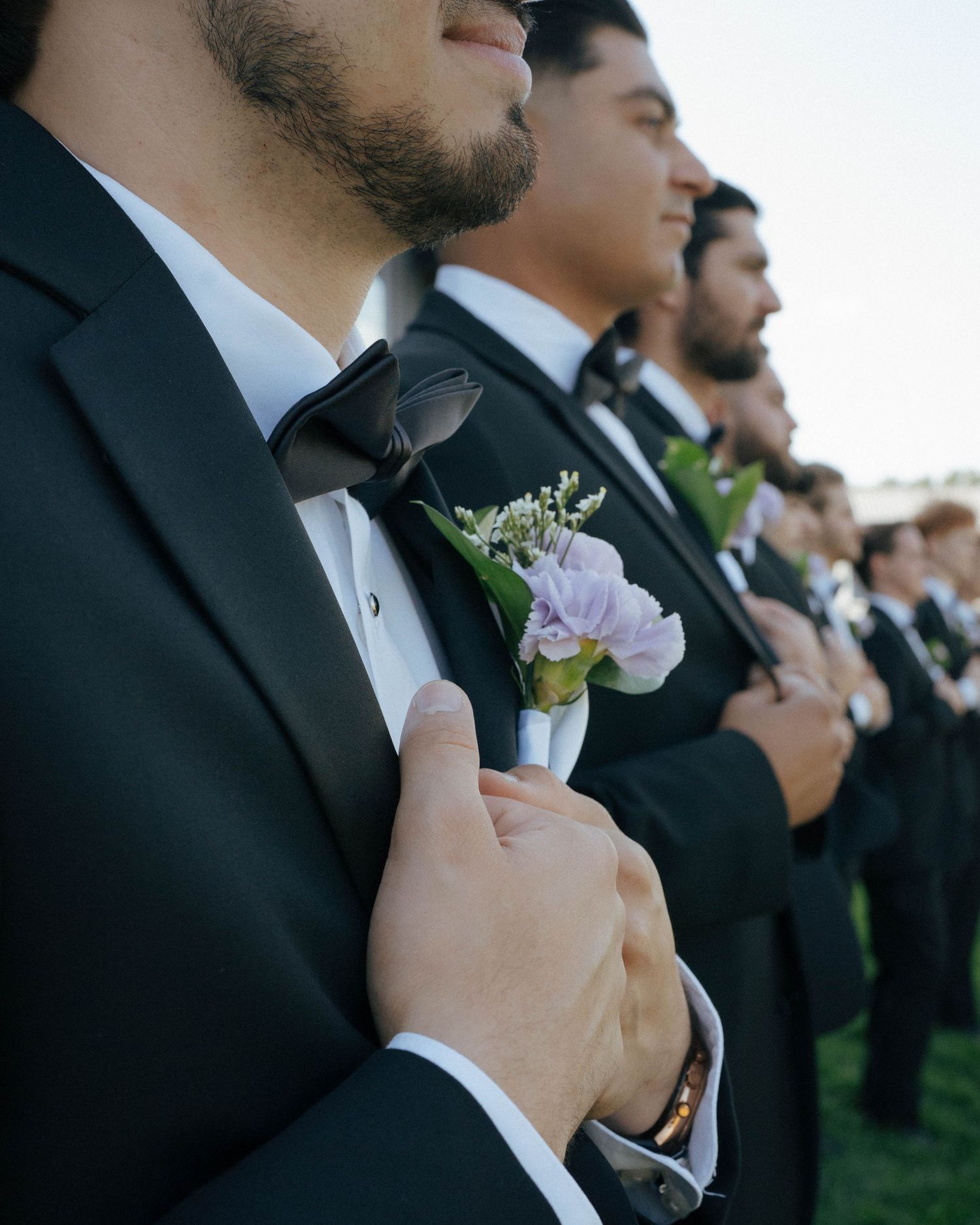 Groomsmen in black tuxedos stand in line wearing delicate purple and white floral boutonnieres.