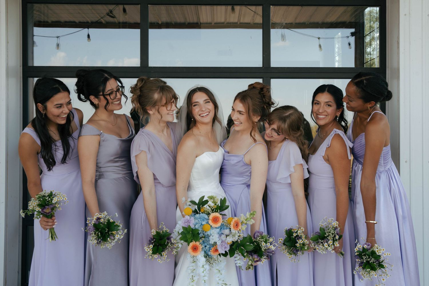 A wedding party in coordinating lavender and purple dresses holds colorful bouquets while posing together.