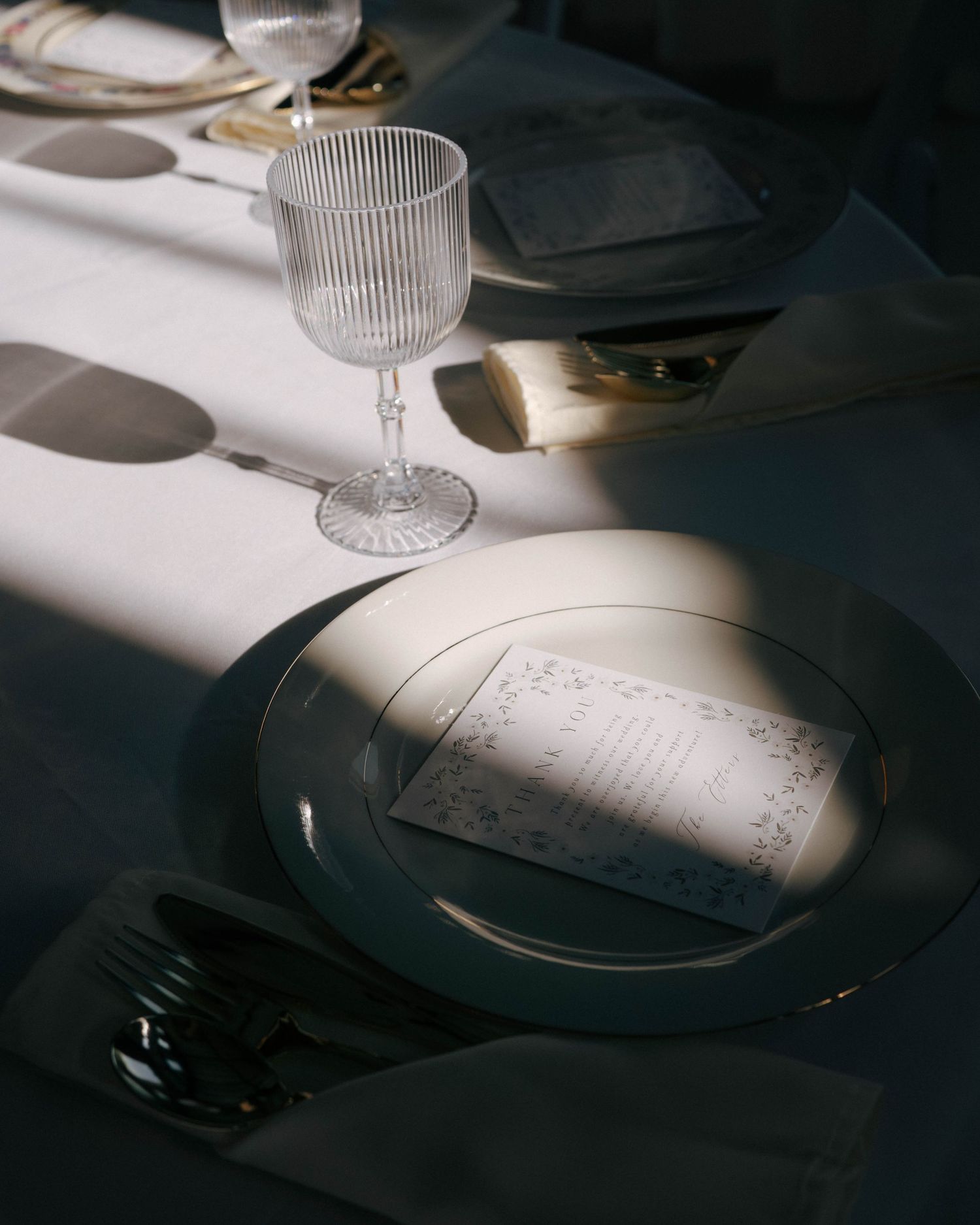 A wine glass and place setting on a white tablecloth casting dramatic shadows in moody lighting.