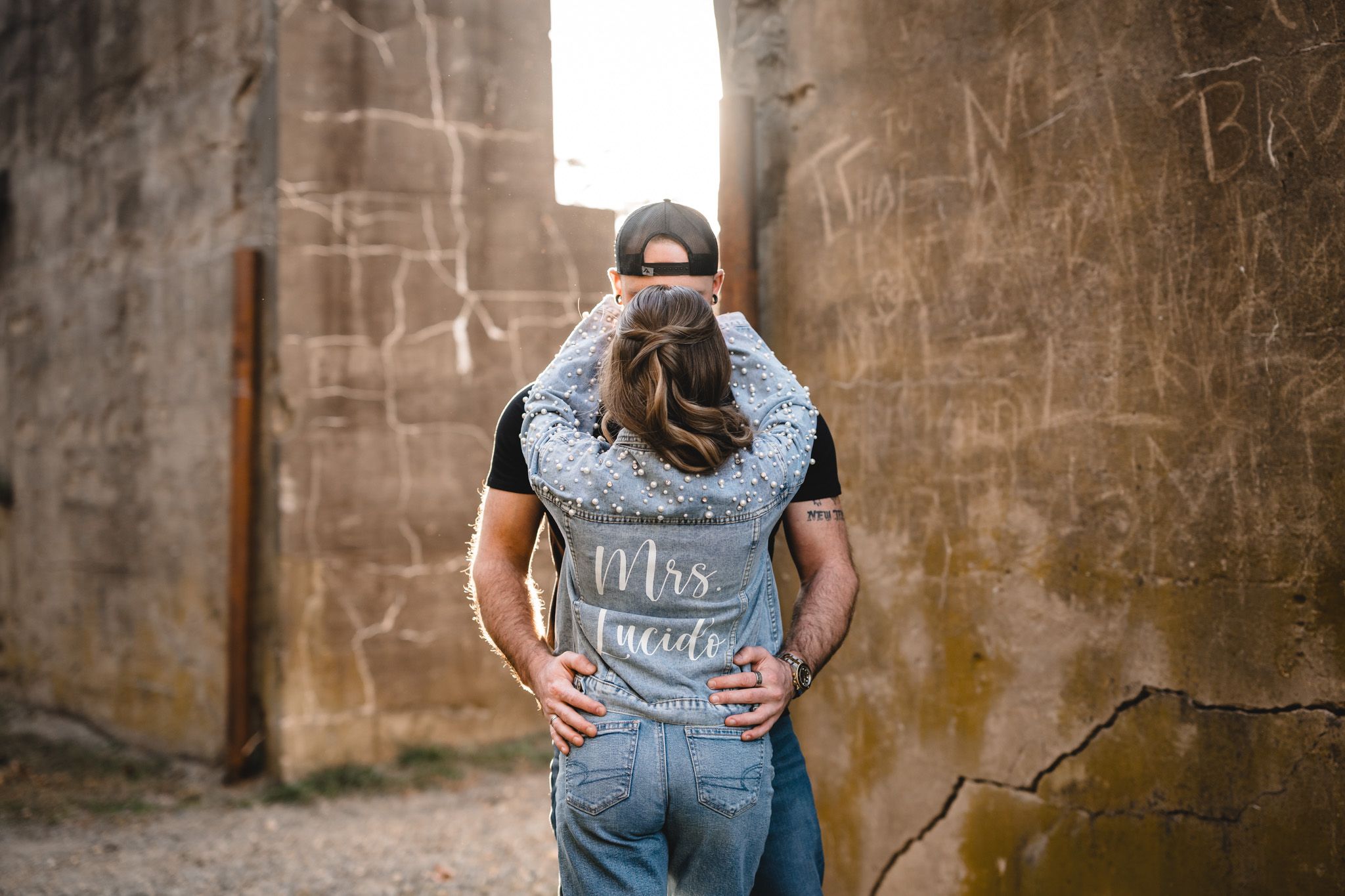A couple embraces against a rustic stone wall in casual denim outfits during an engagement photoshoot.