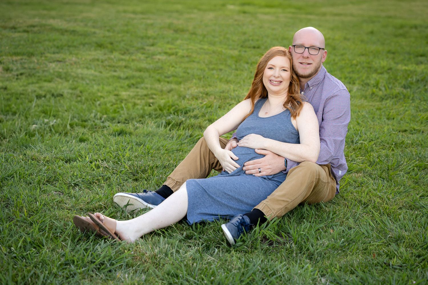 A couple relaxes together on lush green grass during a maternity outdoor photoshoot