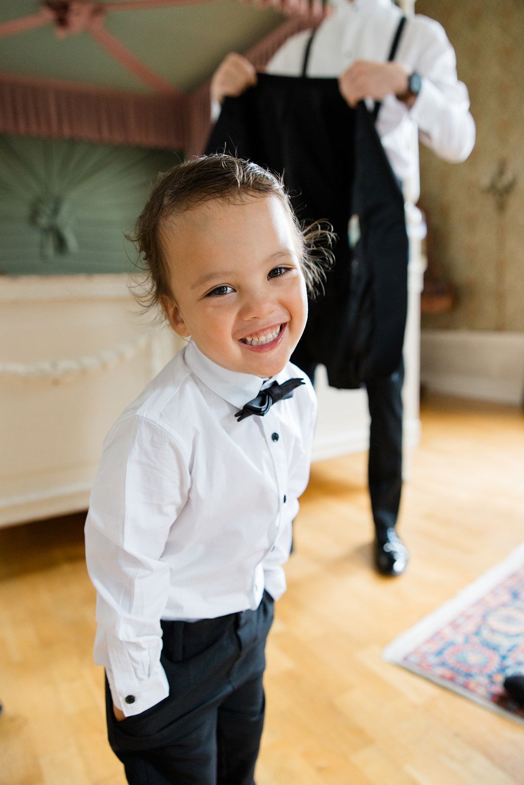 A young child in formal black and white attire smiles while standing on a wooden floor at an indoor event.