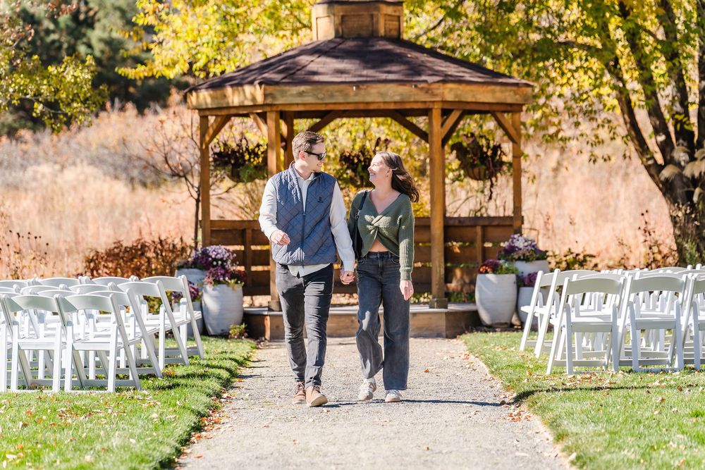 White wedding chairs line a scenic outdoor ceremony venue with fall foliage and a wooden gazebo.