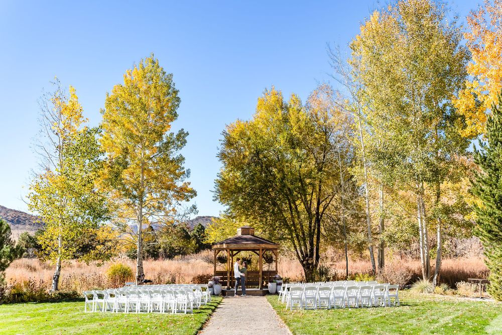 A panoramic view of an outdoor wedding venue with white chairs set up beneath golden autumn trees.